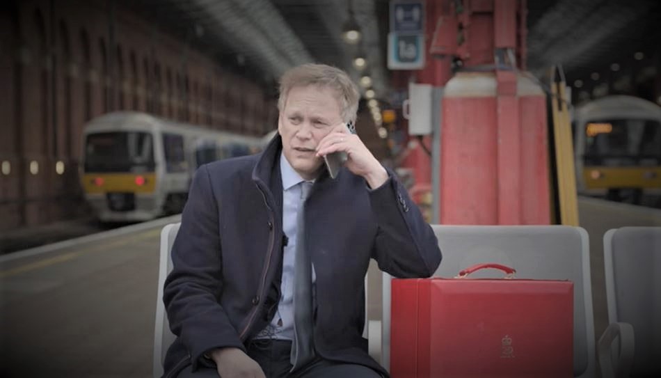 Still image from Grant Shapps' 'Great British Railways Headquarters Competition' video. The colours in the image are dark, suggesting he is having a suspicious phone call on a railway platform.